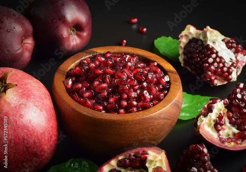 Wooden mortar full ripe arils of pomegranate, red apples, green leaves on black background. Still life fruits, sweet seeds. Split open clusters of pomegranates. Healthy food. Studio shot photo
