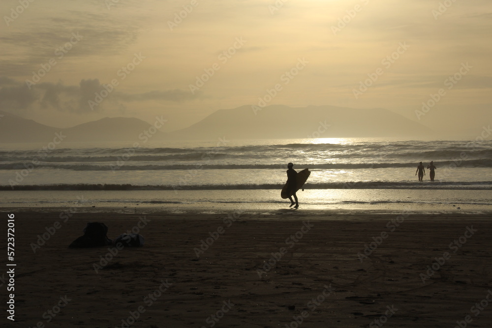 Silhouette of a person surfing at Inch beach at sunset with swimmers in the water (Dingle Peninsula, County Kerry, Ireland).Concept for cold water swimming in winter