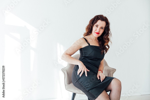 brunette in black dress sits in a chair in a bright room