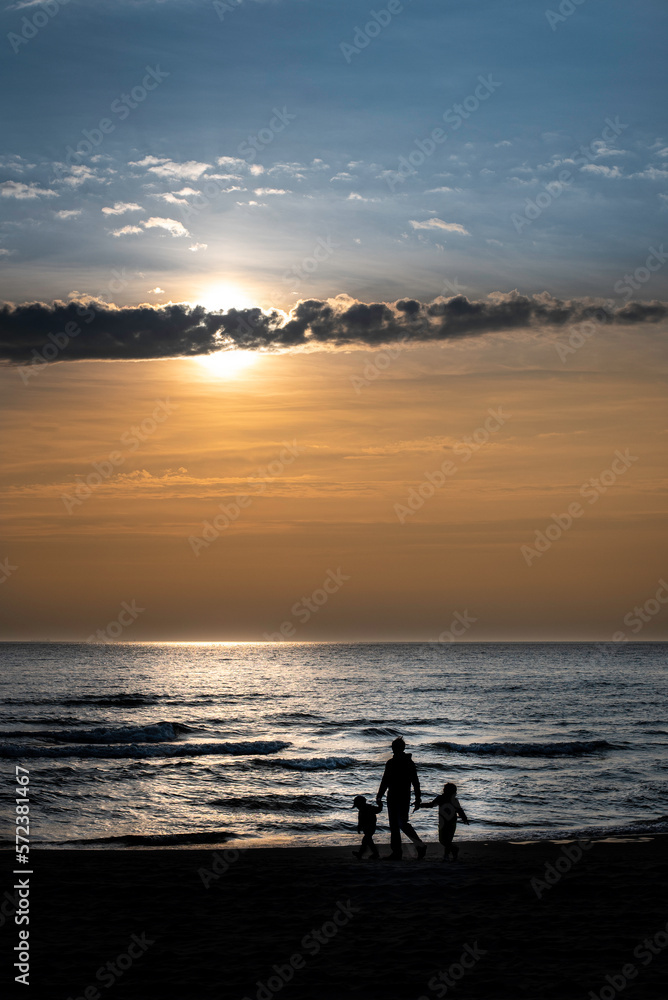 A family of three (parent with 2 children) walking by the sea during a very beautiful yellow sunset