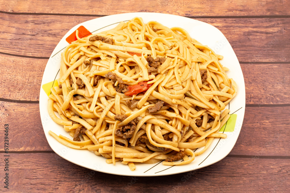 Salted noodles with vegetables, onion and pieces of beef garnished on a white plate on a wooden table Salted noodles with vegetables, onion and pieces of beef garnished on a white plate on a wooden table