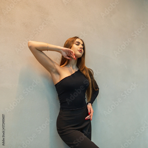 Beautiful elegant young girl in fashionable black clothes with trousers and a one-shoulder top posing near a gray wall