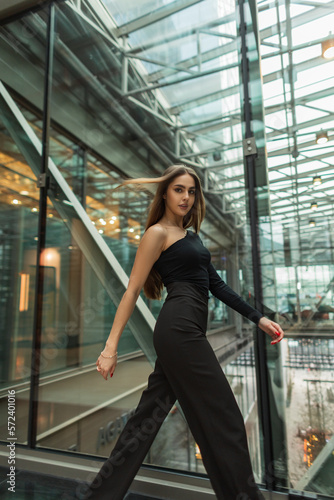 Stylish beautiful young business manager girl in a fashion black clothes with a top and pants is walking in a modern glass office building. Pretty woman professional in motion indoors