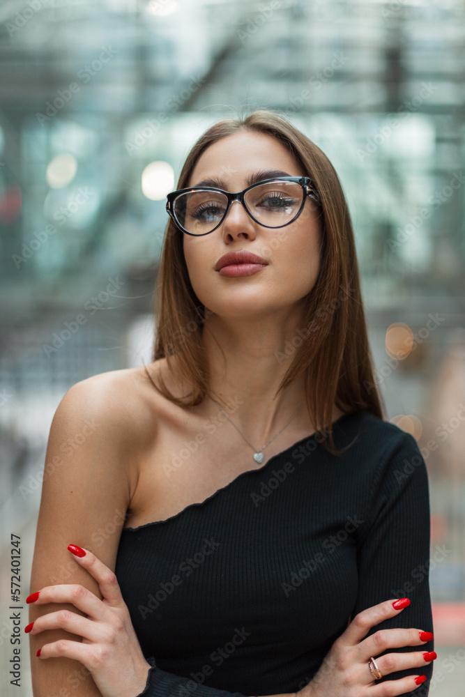 Pretty young specialist professional woman with trendy glasses in a fashionable black top in a glass office building at work. Business beauty girl