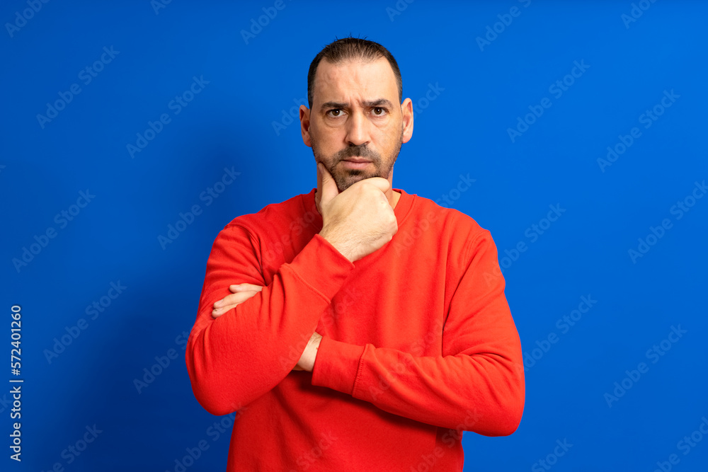 Bearded hispanic man wearing red sweater pensive thinking looking at copyspace hold hand chin isolated over blue color background. Bearded hispanic man wearing red sweater pensive thinking looking at copyspace hold hand chin isolated over blue color background.