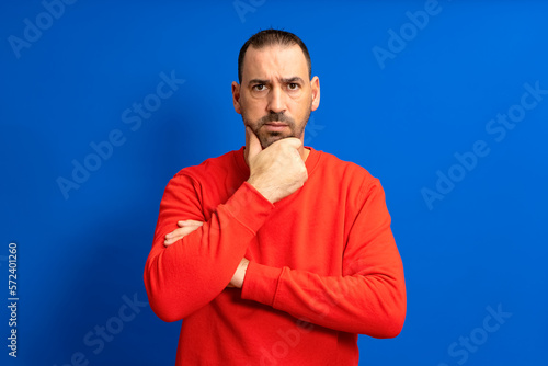 Bearded hispanic man wearing red sweater pensive thinking looking at copyspace hold hand chin isolated over blue color background. © Andres