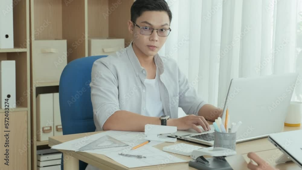 Tilt up portrait shot of young Asian man working on laptop at office desk and then posing for camera Tilt up portrait shot of young Asian man working on laptop at office desk and then posing for camera