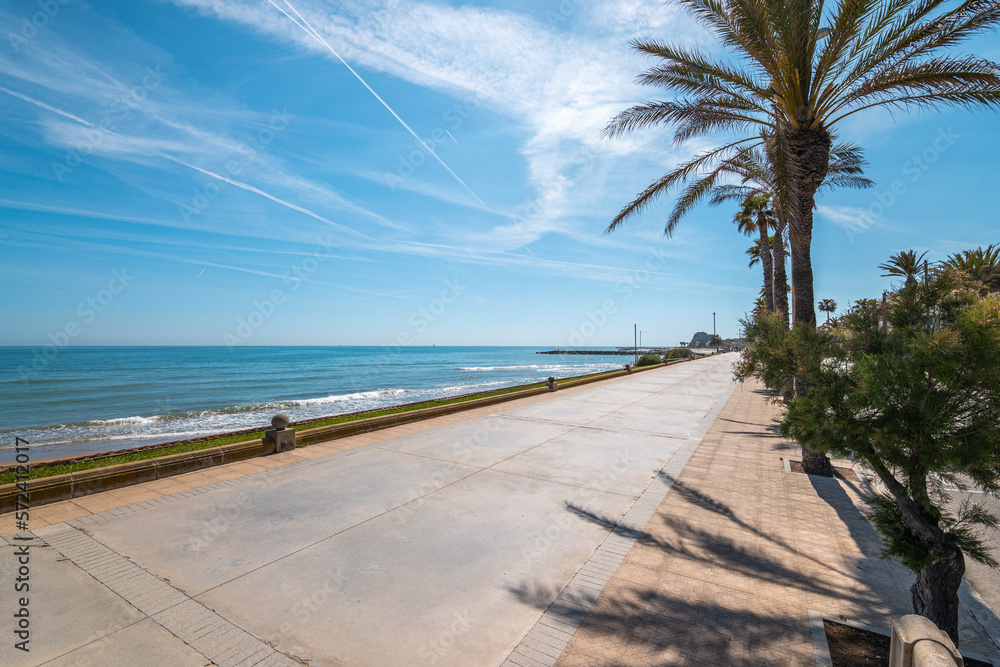 Excellent walks along the shore of the sea with azure water along a pedestrian road made of concrete slabs. Cloudless bright blue sky with bright sunlight overhead. Palm trees grow along the road.