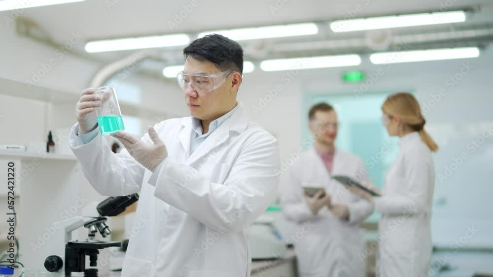 Medical researcher scientist asian man with protective glasses exploring looking at blue chemical liquid in test tube with colleagues at the background at microbiology pharmaceutical biochemistry lab Medical researcher scientist asian man with protective glasses exploring looking at blue chemical liquid in test tube with colleagues at the background at microbiology pharmaceutical biochemistry lab