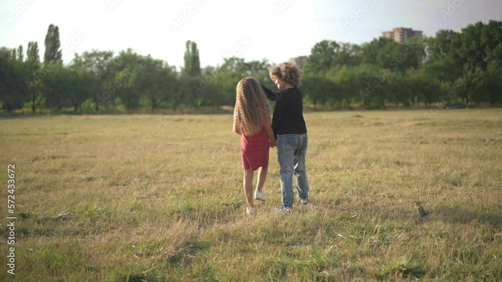 Wide shot carefree happy children walking on sunny spring summer meadow holding hands. Back view relaxed Caucasian boy and girl strolling outdoors at sunset enjoying weekend Wide shot carefree happy children walking on sunny spring summer meadow holding hands. Back view relaxed Caucasian boy and girl strolling outdoors at sunset enjoying weekend
