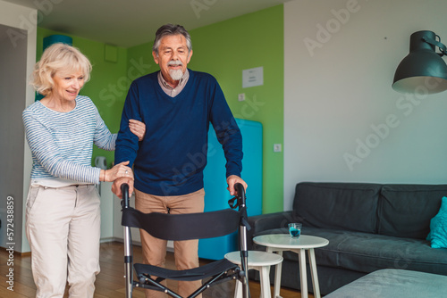 Happy senior couple living together in nursing home, woman helping her husband to walk inside the apartment