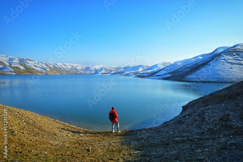 Traveler, a man on the shore of a mountain lake in a red jacket with a backpack, in the background are snowy mountains, hills and a blue sky