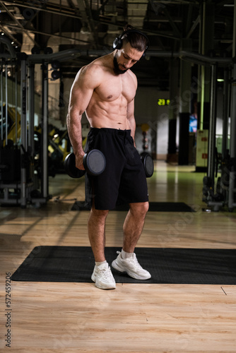 A muscular man with headphones does an exercise with dumbbells dumbbells in the gym