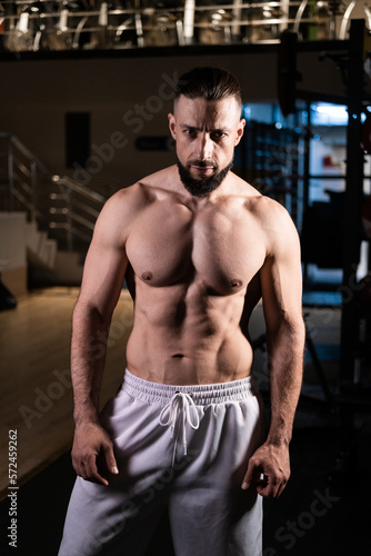 A muscular man with a beard poses in the gym in white pants