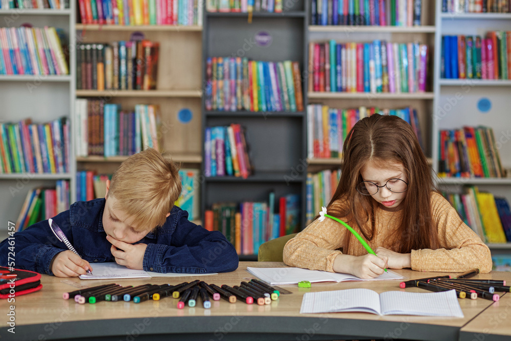 Two schoolchildren write and draw sitting at table in library. Classmates study in classroom. Two schoolchildren write and draw sitting at table in library. Classmates study in classroom.