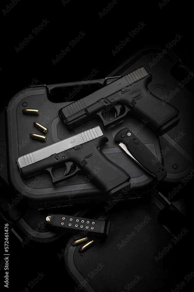 Two pistols, 9mm cartridges and a penknife lie on a plastic gun case. Self-defense and survival kit. Compact edged weapons and firearms. Dark back.