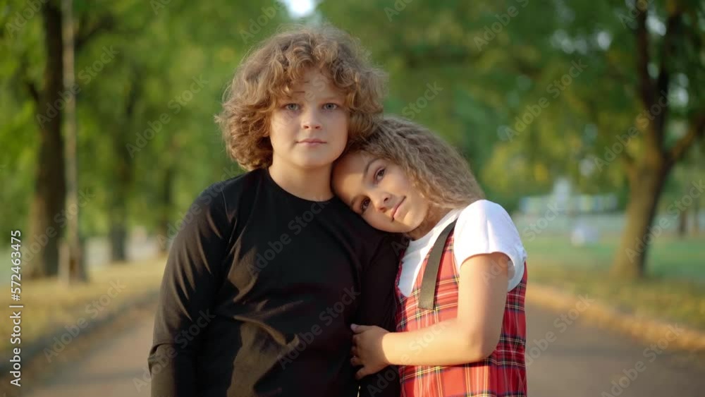 Loving girl hugging boy talking looking at camera standing on sunny summer spring day on park alley. Medium shot portrait of confident Caucasian girlfriend posing dating with boyfriend outdoors Loving girl hugging boy talking looking at camera standing on sunny summer spring day on park alley. Medium shot portrait of confident Caucasian girlfriend posing dating with boyfriend outdoors