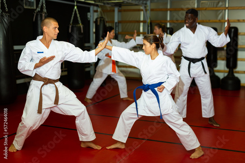 Man and women in kimono sparring together in gym during group karate training. African-american man trainer standing nearby and observing..