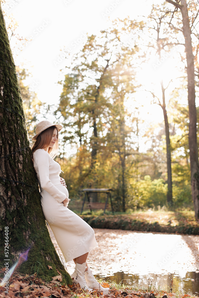 Naklejka premium Elegant young pregnant woman in white knit dress and hat touching and stroking belly, relaxing in park by the lake on autumn warm day. Preparation for childbirth, healthy motherhood. Mothers day