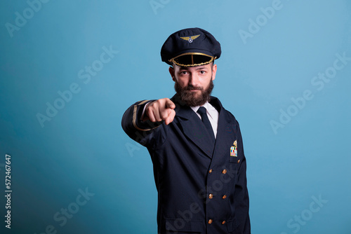 Serious airplane pilot pointing at camera, plane captain wearing uniform and hat front view portrait. Aviation academy aviator with airline wings badge on jacket looking at you photo