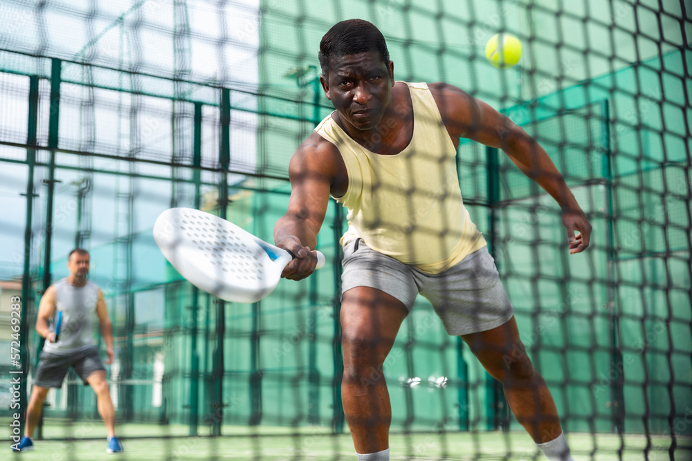 Young adult man playing paddle tennis doubles match with male partner outdoors