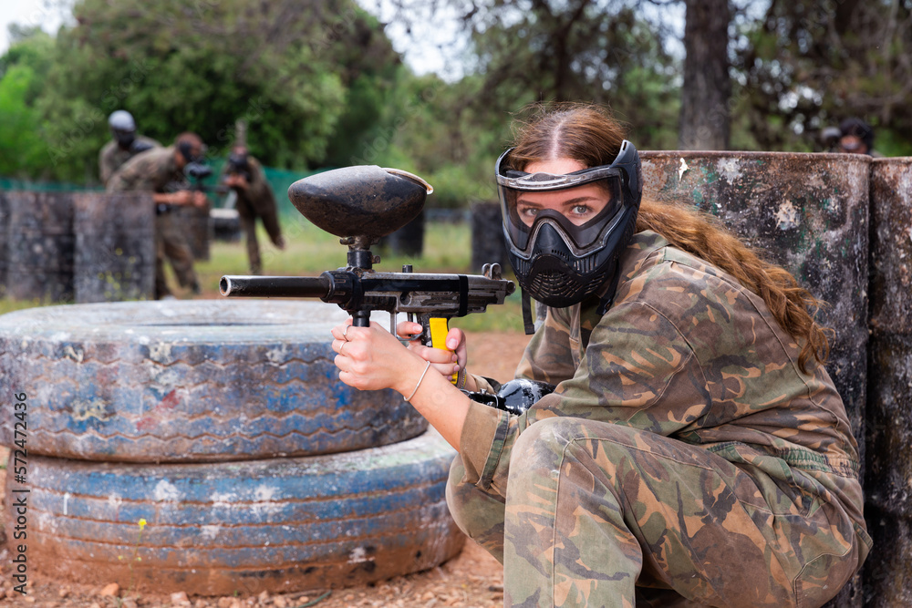 Fototapeta premium Female paintball player in protective mask with ammunition and gun on paintball field