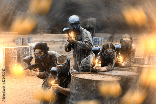 Five paintball players in camouflage and protective masks aims with guns outdoors