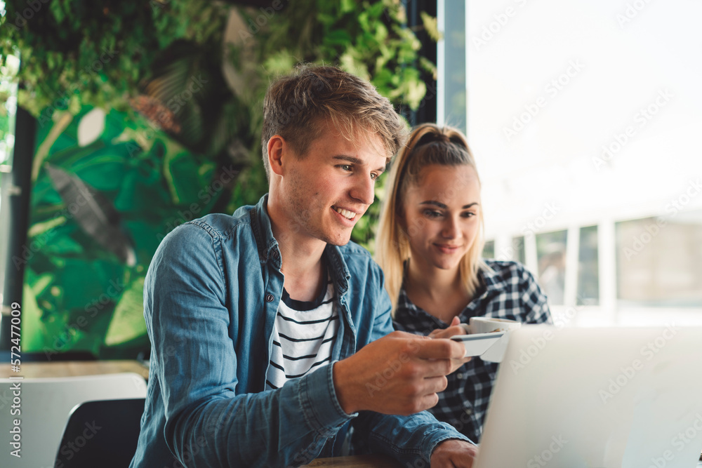 Smiling young man holding a credit card while his girlfriend sitting next to him, finishes the online payment on laptop Smiling young man holding a credit card while his girlfriend sitting next to him, finishes the online payment on laptop