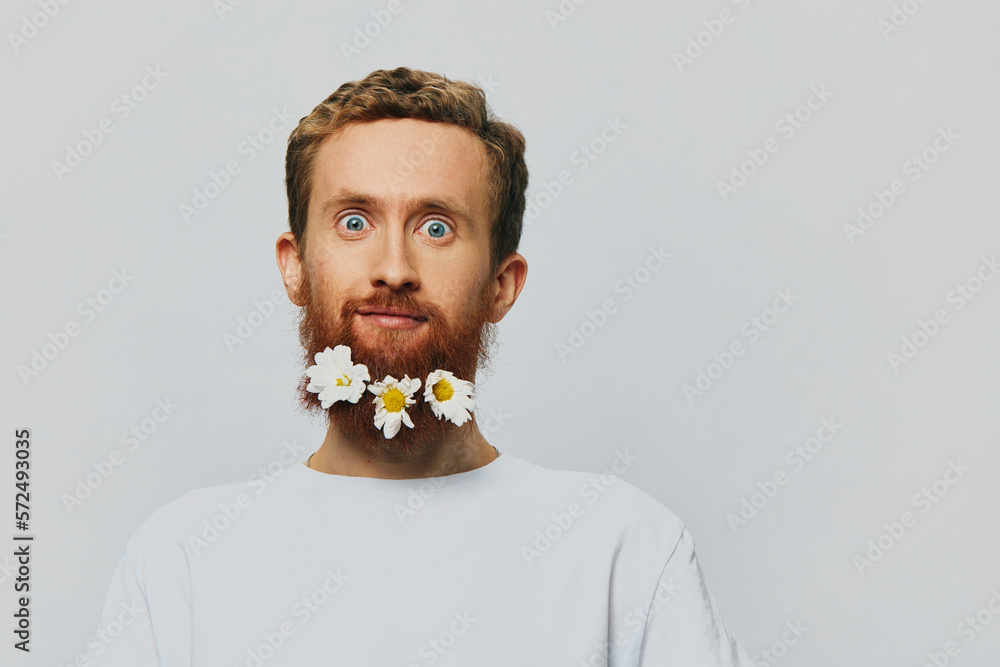 Portrait of a funny man in a white T-shirt with flowers daisies in his beard on a white isolated background, copy place. Holiday concept and congratulations. Portrait of a funny man in a white T-shirt with flowers daisies in his beard on a white isolated background, copy place. Holiday concept and congratulations.