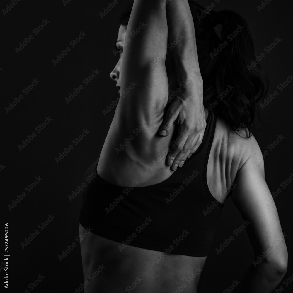 Female sporty muscular with ponytail doing stretching workout of the shoulders, blades in sport bra, holing the neck the hand on dark grey background with empty copy space. Back view. Black and white 