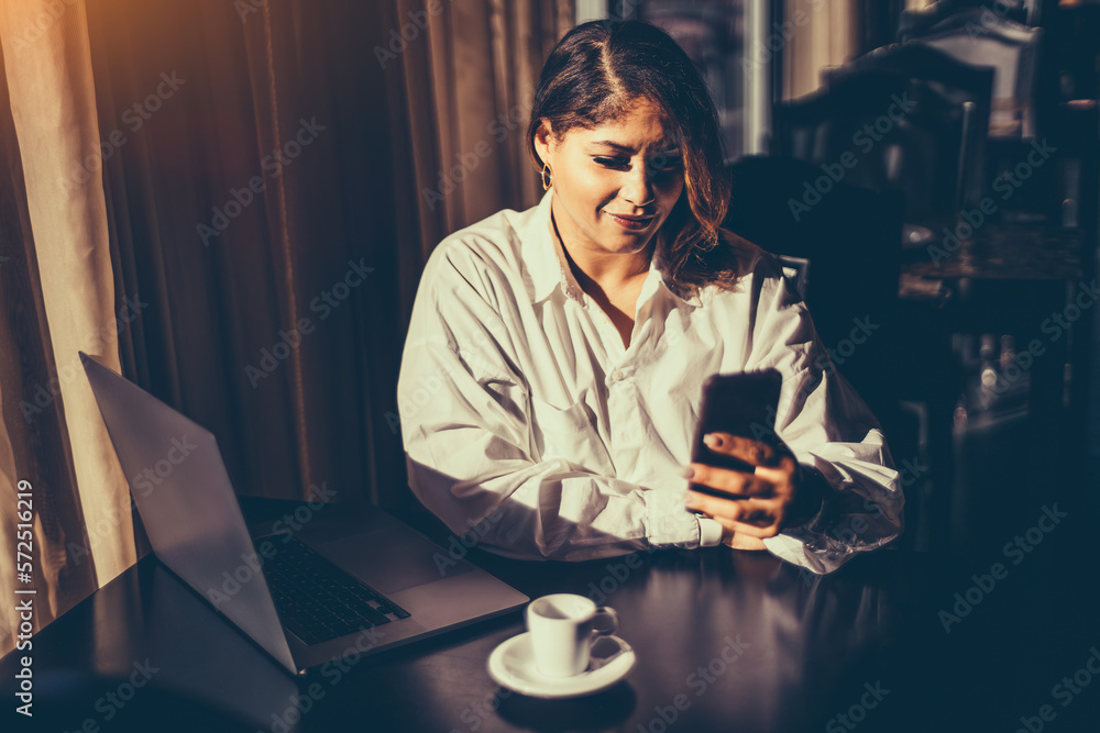 Portrait of an attractive Hispanic woman with a white oversize shirt, receiving sun rays through the coffee shop window while on her cell phone, with her laptop open and her coffee cup at her side Portrait of an attractive Hispanic woman with a white oversize shirt, receiving sun rays through the coffee shop window while on her cell phone, with her laptop open and her coffee cup at her side
