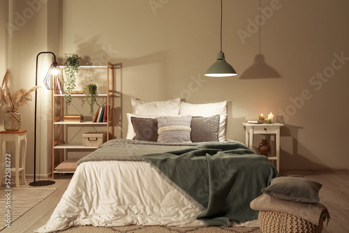 Interior of bedroom with green blankets on bed, burning candles and glowing lamps late in evening