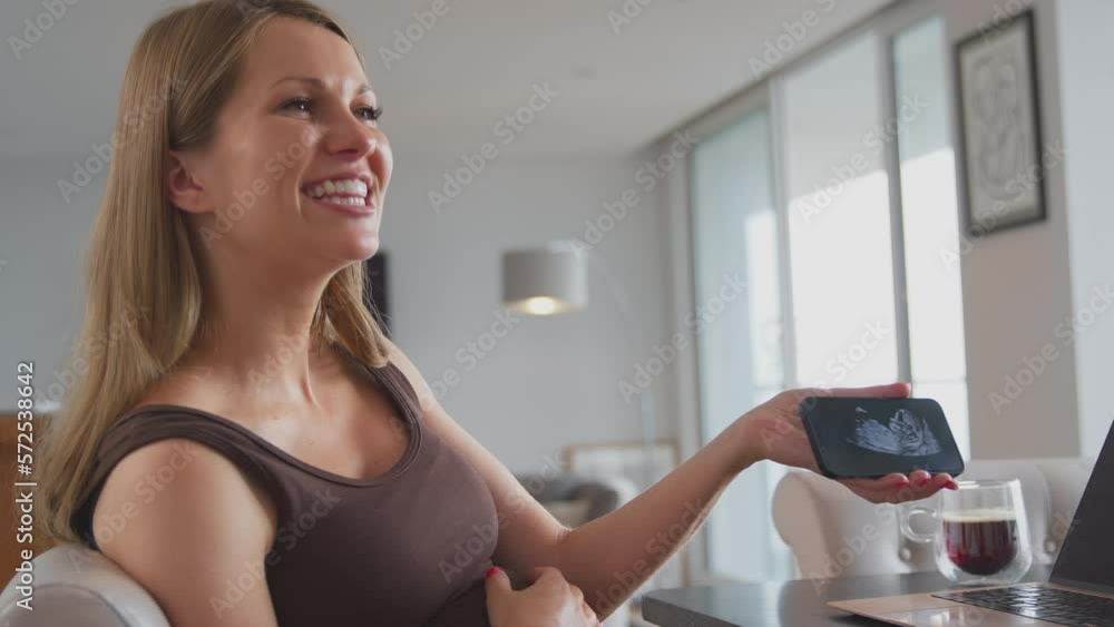 Smiling pregnant woman with hand on stomach at home showing ultrasound scan of baby on phone to visiting off camera friend - shot in slow motion Smiling pregnant woman with hand on stomach at home showing ultrasound scan of baby on phone to visiting off camera friend - shot in slow motion