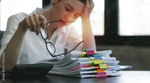 Stressed and tired woman holding glasses while workign on pile of paperwork at office. photo