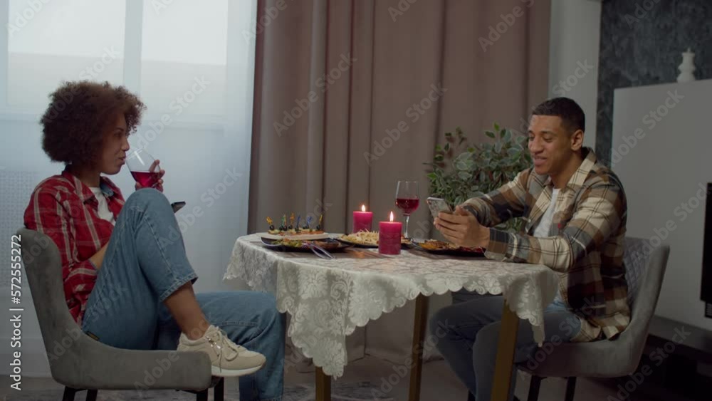 Positive attractive African American man networking on smartphone and talking with pretty black woman, enjoying romantic dinner and bonding while happy couple relaxing in domestic room. Positive attractive African American man networking on smartphone and talking with pretty black woman, enjoying romantic dinner and bonding while happy couple relaxing in domestic room.