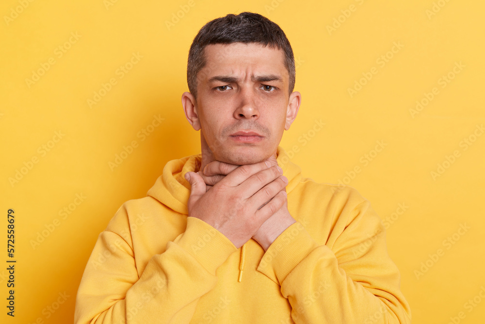 Portrait of ill man with dark hair wearing casual style hoodie standing isolated over yellow background, touching his neck, suffering sore throat, viral infection or flu symptoms. Portrait of ill man with dark hair wearing casual style hoodie standing isolated over yellow background, touching his neck, suffering sore throat, viral infection or flu symptoms.