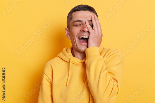 Happy positive overjoyed man wearing casual style hoodie, laughing happily at something keeps hands on face, smiles broadly, expressing positive emotions, standing isolated over yellow background.