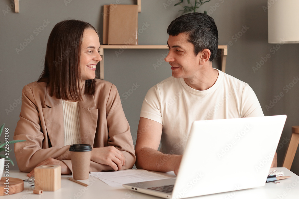Portrait of cheerful young Caucasian woman and man using laptop sitting at table with notebook in modern office, looking at each other, smiling, working together. Portrait of cheerful young Caucasian woman and man using laptop sitting at table with notebook in modern office, looking at each other, smiling, working together.