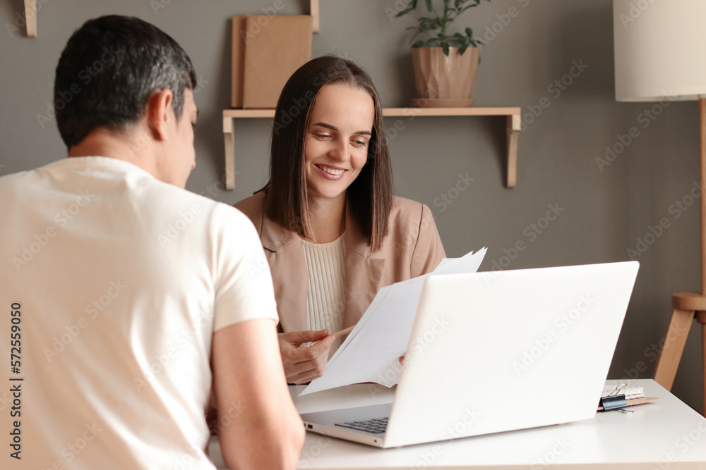 Indoor shot of smiling businesswoman working with new partner, showing documents, sitting at table in office in front of laptop, colleagues negotiating a new contract. Indoor shot of smiling businesswoman working with new partner, showing documents, sitting at table in office in front of laptop, colleagues negotiating a new contract.