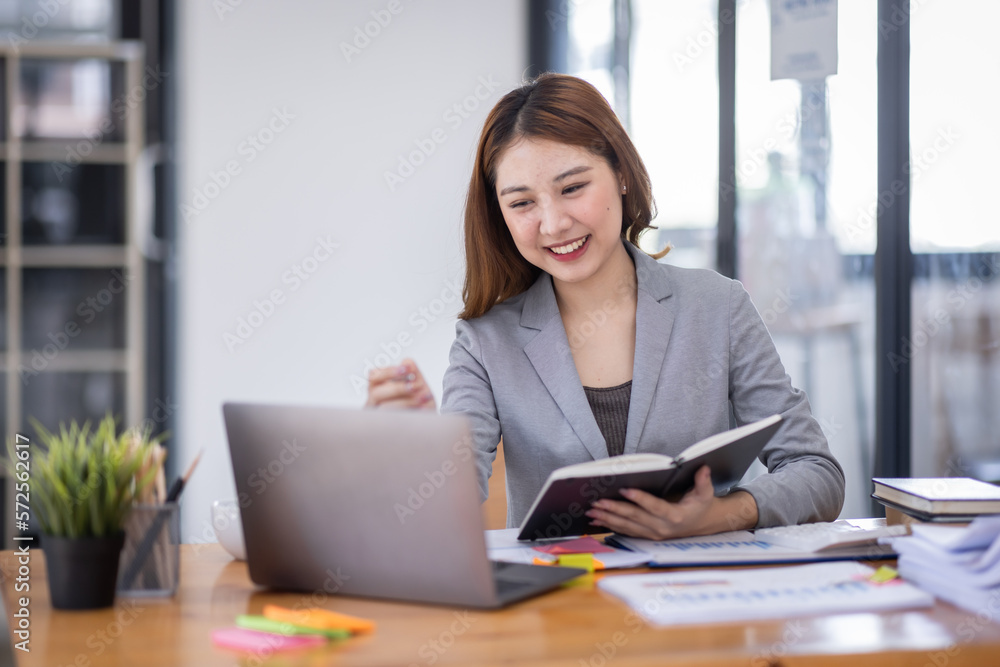 Business Asian woman using calculator and writing make note with calculate doing math finance on an office desk. Woman working at office with laptop and tax, accounting, documents on desk.