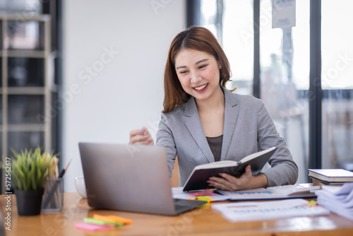 Business Asian woman using calculator and writing make note with calculate doing math finance on an office desk. Woman working at office with laptop and tax, accounting, documents on desk.