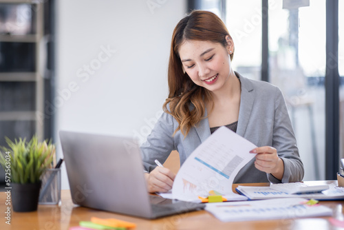 Business Asian woman using calculator and writing make note with calculate doing math finance on an office desk. Woman working at office with laptop and tax, accounting, documents on desk.