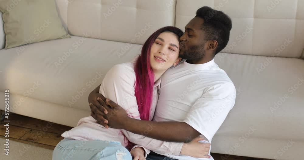 Black guy hugs his girlfriend, a caucasian young woman with colored hair. A loving multiracial married couple is sitting near the comfortable sofa in the cozy living room. Multinational relationship. Black guy hugs his girlfriend, a caucasian young woman with colored hair. A loving multiracial married couple is sitting near the comfortable sofa in the cozy living room. Multinational relationship.