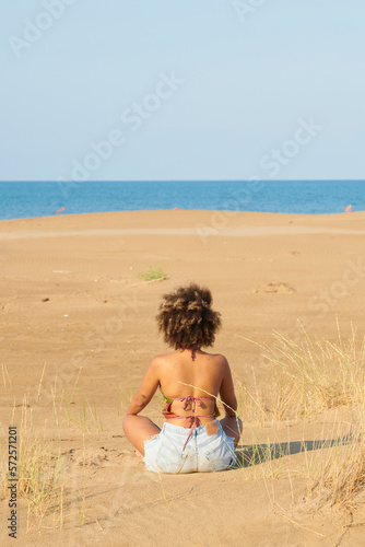Woman meditating looking at endless sea from hill. Back view of frizzy-haired woman in sleeveless top and denim shorts sitting on sandy slope and watching idyllic sea on clear sunny day.