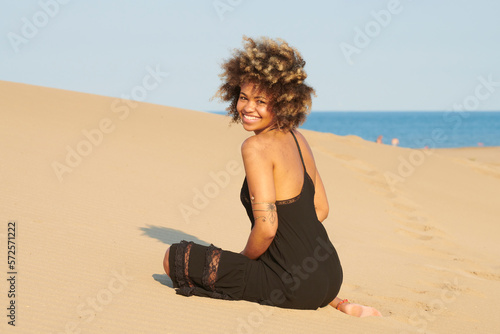 Attractive joyful woman with frizzy hair in black dress smiling at camera over shoulder at the sea. Back view of pretty delighted woman with frizzy hair wearing summer black dress sitting on the sand.