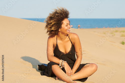 Entertained woman with frizzy hair in black summer dress laughing on the beach. Portrait of woman with frizzy hair in the wind having fun on sandy beach on summer day. 