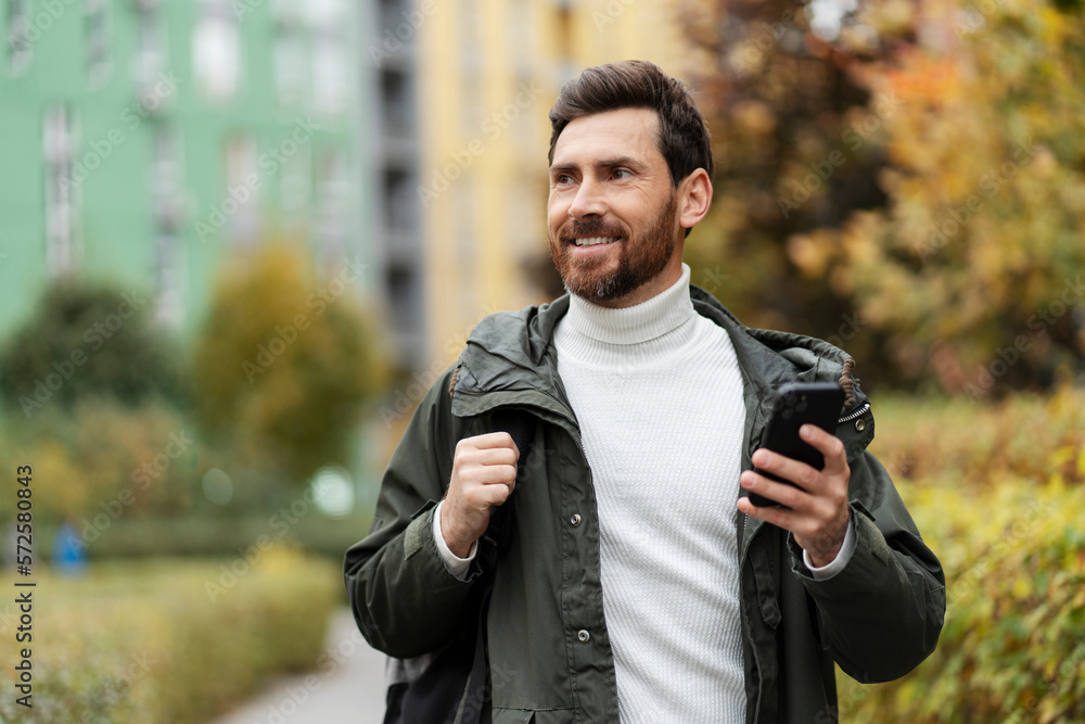 Handsome man with a beard on the city street, holding a phone in his hand. He has a backpack on his shoulder, he looks away with a smile. Handsome man with a beard on the city street, holding a phone in his hand. He has a backpack on his shoulder, he looks away with a smile.
