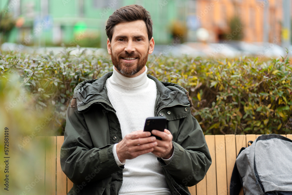 Bearded man holds a phone in his hands, looks at the camera, smiles sweetly. A man outside, sitting on a bench. Bearded man holds a phone in his hands, looks at the camera, smiles sweetly. A man outside, sitting on a bench.