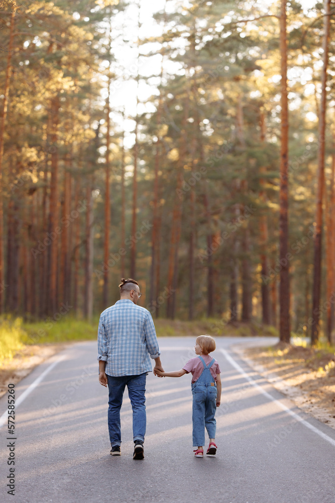 Fototapeta premium Dad and his little daughter are walking along a forest road among tall pines. Family walk in the forest at sunset, man and little girl.