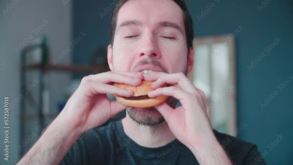 Slow motion young caucasian man bites into burger at home, cheeseburger eating enjoyment, close up portrait fast food eating, unhealthy eating concept obesity Slow motion young caucasian man bites into burger at home, cheeseburger eating enjoyment, close up portrait fast food eating, unhealthy eating concept obesity